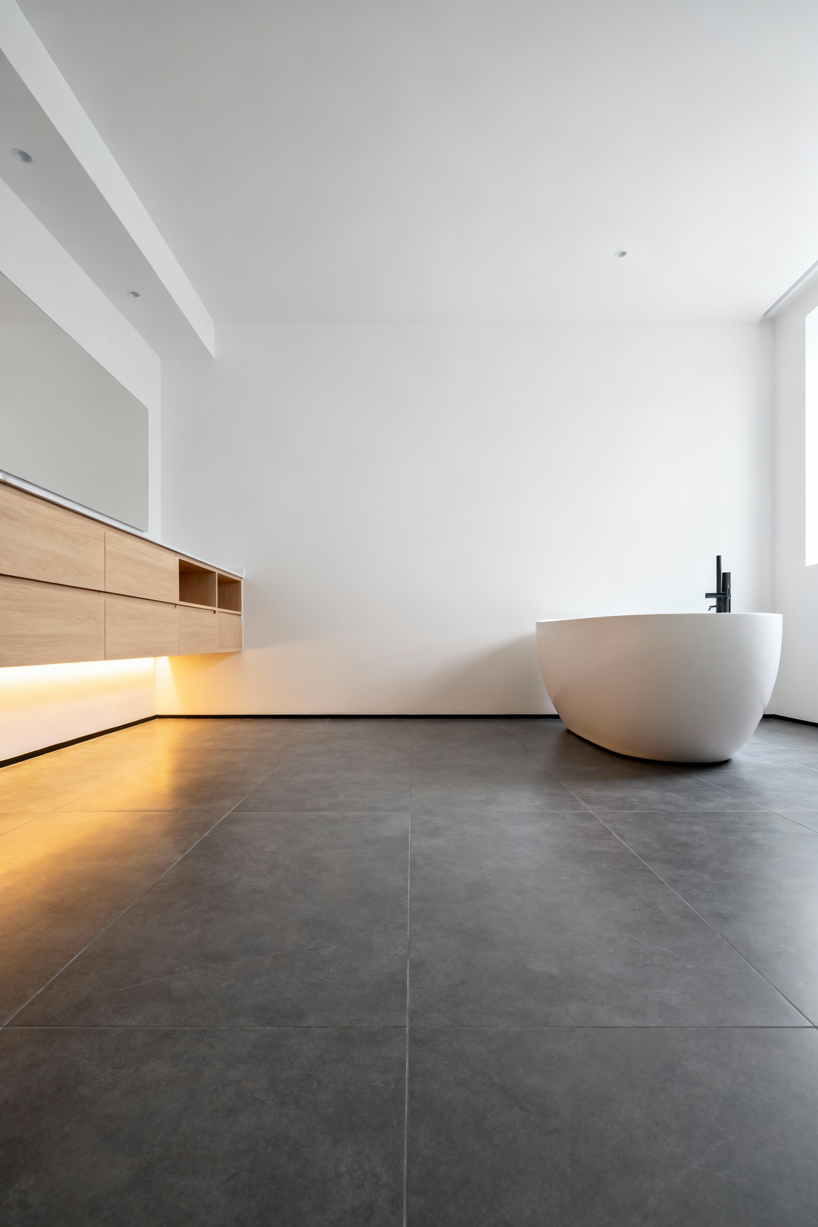 Luxury minimalist bathroom interior featuring seamless slate-gray porcelain floor tiles and a freestanding tub, illuminated by warm light suggesting hidden radiant heating systems.
