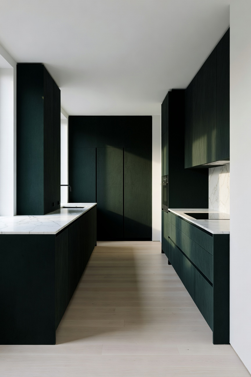 A modern minimalist kitchen with deep forest green cabinets, white walls, and light wood flooring illuminated by natural sunlight.