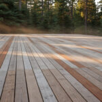 A wide cedar deck showing a weathered silvery-gray patina transitioning from the original warm reddish-brown wood color, set in a natural forest environment.